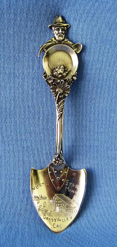 Souvenir Mining Spoon North Star  Mine.JPG - SOUVENIR MINING SPOON NORTH STAR MINE - Sterling silver spoon engravedwith a detailed picture of the North Star Mine Grass Valley, Cal; miner figure with gold pan and crossed shovel and pick on handle, 4 in. long, weight 11 gms., marked on back Sterling with a mfg. hallmark  (The North Star Mine was located on Lafayette Hill a short distance south of Grass Valley, CA. The Grass Valley District was the second largest producer of gold during California’s Gold Rush behind the Mother Lode of the Sierra Nevada district. The Lafayette ledge by Wolf Creek was worked since 1851, and was pronounced by the State Geologist in 1855 as being one of the best-producing for quartz mining in California.  It was discovered during the time of the California Gold Rush by a group of men, principally Frenchmen, who named their company, the "Helvetia and Lafayette Company". It changed hands in 1855, and again in 1857 when it was purchased under a forced sale for US$15,000. At the same time the name was changed to the "North Star." In the 1860s, reserves were estimated to be not less than thirty thousand tons, worth in the aggregate of $900,000.  Competition between Grass Valley Gold District's 95 mines was fierce, forcing them to open, close, and re-open at various times. The North Star Mine was Grass Valley Gold District's deepest mine, measuring 4,000 feet vertical depth.  In 1895 the mine owners hired A. D. Foote to design and construct an electric-generating plant for the mine.  A 30-foot diameter Pelton water wheel, the largest in the world to that date, was installed in the North Star Mine powerhouse.  By 1928, the North Star's total output value was approximately $33 million.  The following year, Newmont Mining Corporation purchased the adjacent Empire Mine and the North Star Mine, consolidating them to become Empire-Star Mines, Ltd. The Empire-Star was forced to shut down by the U.S. War Production Board during World War II.  Mining resumed after the war but inflation costs of gold mining left the operation unprofitable.  The mine was officially closed on May 28, 1957 when the last water pumps were shut down and removed.  In 1975 California State Parks purchased the Empire-Star property to create a state historic park.  The North Star powerhouse is designated as a California Historical Landmark and serves today as a wonderful museum featuring the Pelton wheel and other relics of the mining past.)